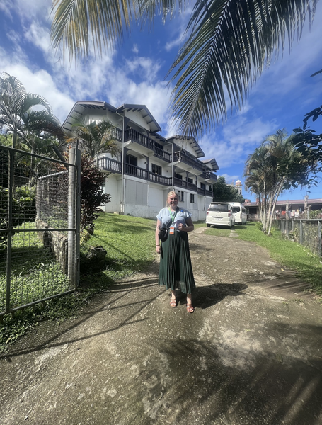 伊莎贝尔·伦德伯格身穿绿色百褶裙和灰色衬衫，站在斐济一座带有棕色阳台的两层白色建筑前